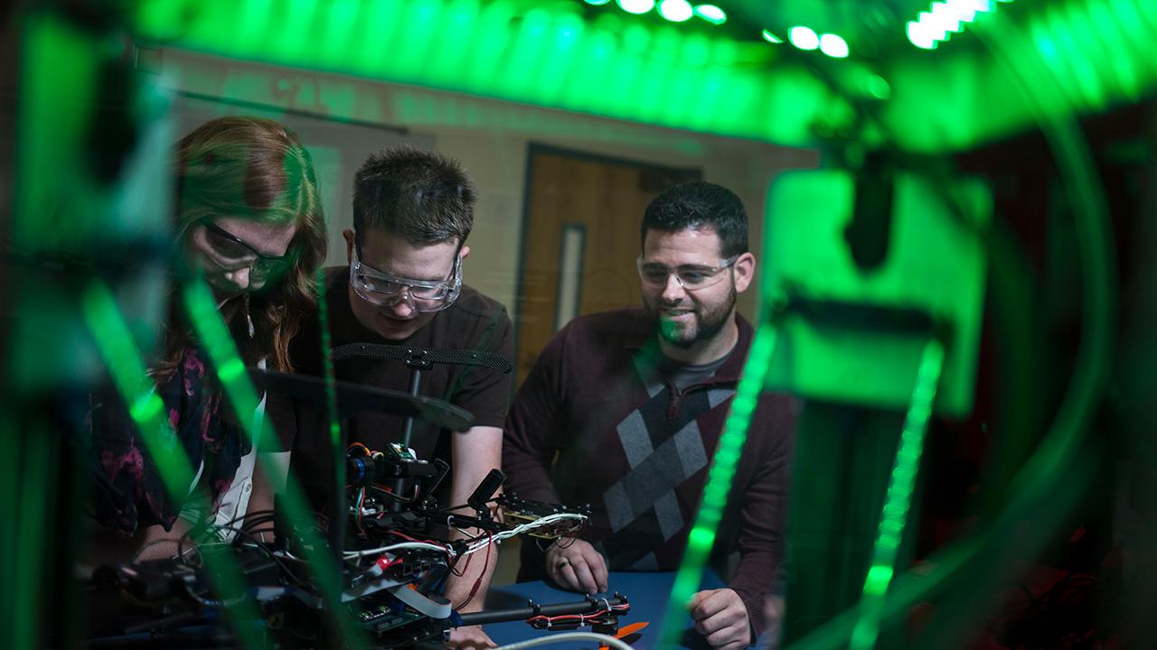 Three students, two male and one female, are working on a drone in an NMT lab space.